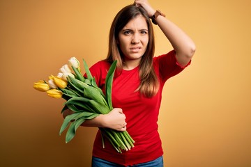 Young blonde woman holding romantic bouquet of tulips flowers over yellow background confuse and wondering about question. Uncertain with doubt, thinking with hand on head. Pensive concept.