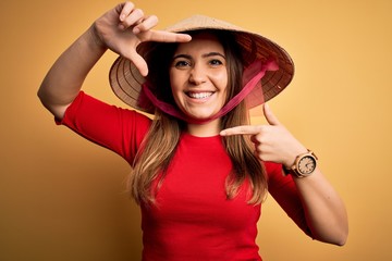 Young blonde woman wearing traditional asian rice paddy straw hat over yellow background smiling making frame with hands and fingers with happy face. Creativity and photography concept.