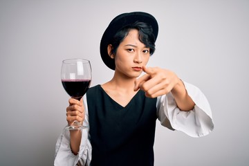 Young beautiful asian sommelier girl drinking glass of red wine over isolated white background pointing with finger to the camera and to you, hand sign, positive and confident gesture from the front