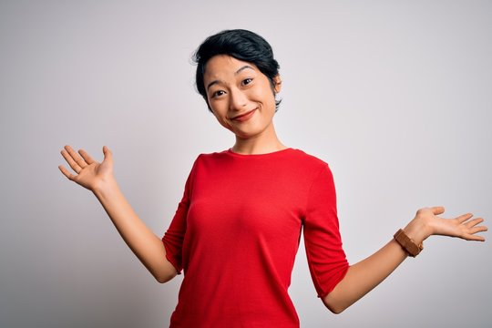 Young beautiful asian girl wearing casual red t-shirt standing over isolated white background clueless and confused expression with arms and hands raised. Doubt concept.