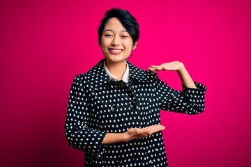 Young beautiful asian girl wearing casual jacket standing over isolated pink background gesturing with hands showing big and large size sign, measure symbol. Smiling looking at the camera. Measuring