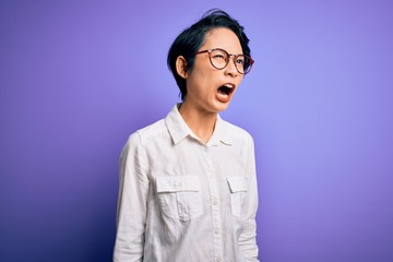 Young beautiful asian girl wearing casual shirt and glasses standing over purple background angry and mad screaming frustrated and furious, shouting with anger. Rage and aggressive concept.