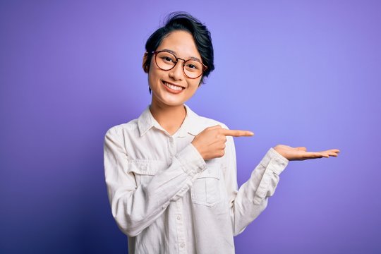 Young beautiful asian girl wearing casual shirt and glasses standing over purple background amazed and smiling to the camera while presenting with hand and pointing with finger.