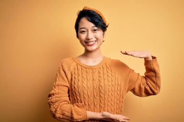Young beautiful asian girl wearing casual sweater and diadem standing over yellow background gesturing with hands showing big and large size sign, measure symbol. Smiling looking at the camera