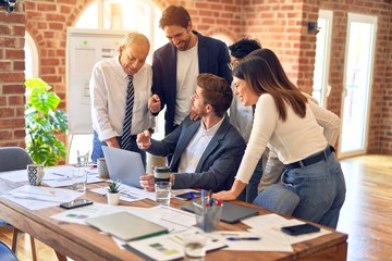 Group of business workers smiling happy and confident. One of them sitting and partners standing around. Working together with smile on face looking at the laptop at the office