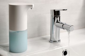 An automatic soap dispenser in the bathroom next to a chrome tap and water pouring from it.