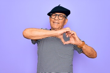 Senior handsome hoary man wearing french beret and glasses standing over purple background smiling in love showing heart symbol and shape with hands. Romantic concept.