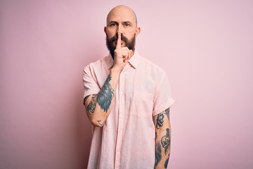 Handsome bald man with beard and tattoo wearing casual shirt over isolated pink background asking to be quiet with finger on lips. Silence and secret concept.