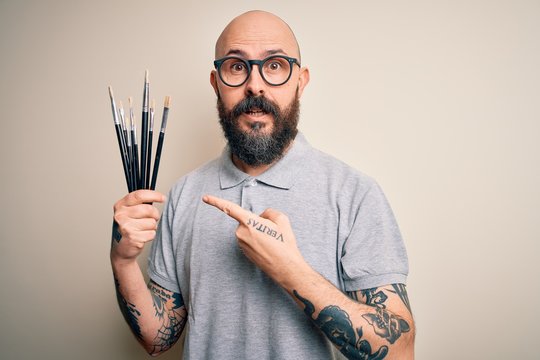 Handsome bald artist man with beard and tattoo painting using painter brushes very happy pointing with hand and finger