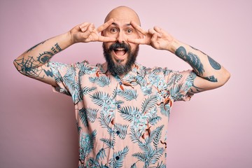 Handsome bald man with beard and tattoo wearing casual floral shirt over pink background Doing peace symbol with fingers over face, smiling cheerful showing victory