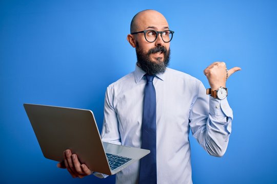 Handsome bald business man with beard working using laptop over blue background pointing and showing with thumb up to the side with happy face smiling