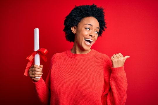 Young African American afro student woman with curly hair holding university degree diploma pointing and showing with thumb up to the side with happy face smiling