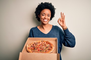 Young African American afro woman with curly hair holding delivery box with Italian pizza doing ok sign with fingers, excellent symbol