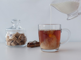 Tea with milk is poured into a glass mug with cinnamon and brown sugar on a light background....