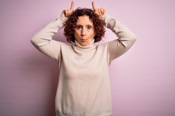 Middle age beautiful curly hair woman wearing casual turtleneck sweater over pink background doing funny gesture with finger over head as bull horns