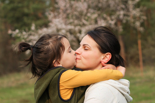 Beautiful mother and daughter hugging and kissing in casual clothes. Warm baby hug on the background of a blurry flowering tree