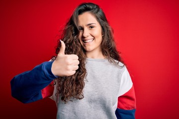 Young beautiful woman with curly hair wearing casual sweatshirt over isolated red background doing happy thumbs up gesture with hand. Approving expression looking at the camera showing success.