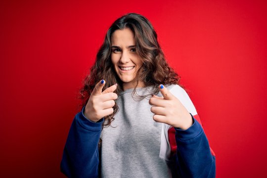 Young beautiful woman with curly hair wearing casual sweatshirt over isolated red background pointing fingers to camera with happy and funny face. Good energy and vibes.
