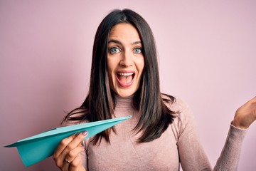 Young woman with blue eyes holding paper airplane standing over isolated pink background very happy and excited, winner expression celebrating victory screaming with big smile and raised hands