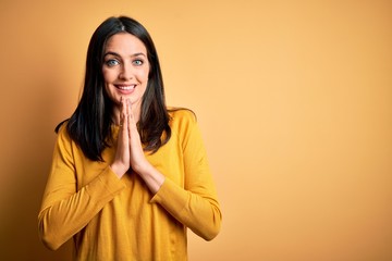 Young brunette woman with blue eyes wearing casual sweater over yellow background praying with hands together asking for forgiveness smiling confident.