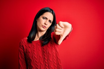 Young brunette woman with blue eyes wearing casual sweater over isolated red background looking unhappy and angry showing rejection and negative with thumbs down gesture. Bad expression.