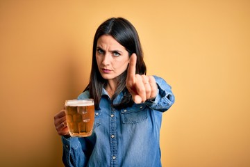 Young woman with blue eyes drinking jar of beer standing over isolated yellow background Pointing with finger up and angry expression, showing no gesture