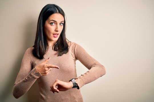 Young brunette woman with blue eyes wearing casual sweater over isolated white background In hurry pointing to watch time, impatience, upset and angry for deadline delay