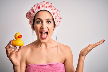 Young brunette woman with blue eyes wearing bath towel and shower cap holding duck toy very happy and excited, winner expression celebrating victory screaming with big smile and raised hands