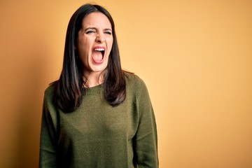 Young brunette woman with blue eyes wearing green casual sweater over yellow background angry and mad screaming frustrated and furious, shouting with anger. Rage and aggressive concept.