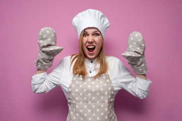 crazy girl cook in uniform shouts and raises hands and shows baking gloves on a colored background,...