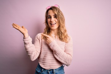 Fototapeta na wymiar Young beautiful blonde woman wearing casual sweater and sunglasses over pink background amazed and smiling to the camera while presenting with hand and pointing with finger.