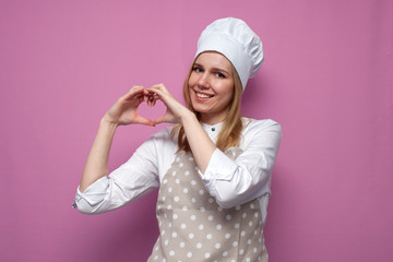 cheerful girl cook in apron shows hands with heart on pink background, woman housewife in kitchen clothes, love of cooking
