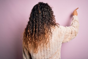 Young beautiful woman with curly hair wearing casual sweater standing over pink background Posing backwards pointing ahead with finger hand