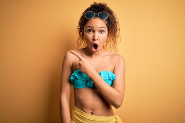 Young beautiful american woman on vacation wearing bikini over isolated yellow background Surprised...