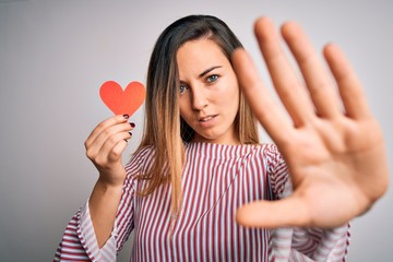 Young beautiful blonde romantic woman holding red paper heart over white background with open hand doing stop sign with serious and confident expression, defense gesture