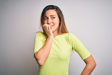 Beautiful blonde woman with blue eyes wearing green casual t-shirt over white background looking stressed and nervous with hands on mouth biting nails. Anxiety problem.