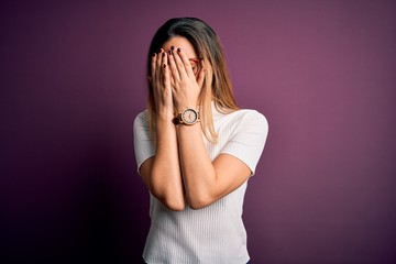 Young beautiful blonde woman with blue eyes wearing casual t-shirt over purple background with sad expression covering face with hands while crying. Depression concept.