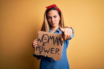 Young beautiful blonde woman with blue eyes asking for women rights holding banner pointing with finger to the camera and to you, hand sign, positive and confident gesture from the front