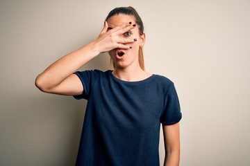 Young beautiful blonde woman with blue eyes wearing casual t-shirt over white background peeking in shock covering face and eyes with hand, looking through fingers with embarrassed expression.