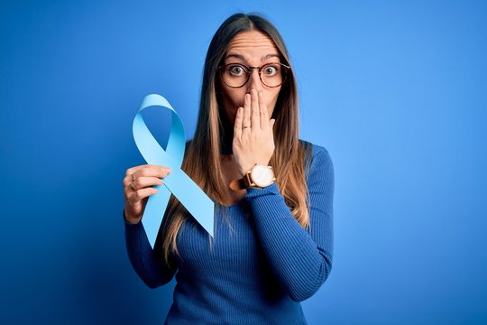 Young Blonde Woman With Blue Eyes Holding Colon Cancer Awareness Blue Ribbon Cover Mouth With Hand Shocked With Shame For Mistake, Expression Of Fear, Scared In Silence, Secret Concept