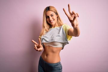 Young beautiful blonde sportswoman doing sport wearing sportswear over pink background smiling looking to the camera showing fingers doing victory sign. Number two.
