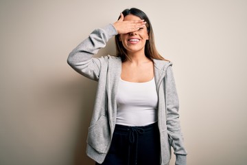 Young beautiful brunette sportswoman wearing sportswoman training over white background smiling and laughing with hand on face covering eyes for surprise. Blind concept.