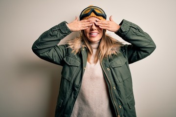 Young brunette skier woman wearing snow clothes and ski goggles over white background covering eyes with hands smiling cheerful and funny. Blind concept.