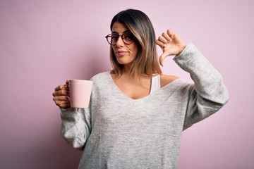 Young beautiful brunette woman wearing glasses drinking pink mug of coffee with angry face,...