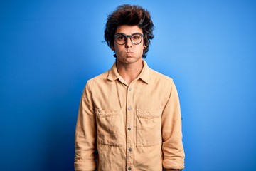 Young handsome man wearing casual shirt standing over isolated blue background puffing cheeks with funny face. Mouth inflated with air, crazy expression.