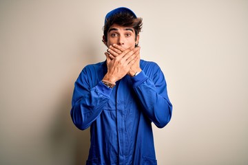 Young mechanic man wearing blue cap and uniform standing over isolated white background shocked covering mouth with hands for mistake. Secret concept.