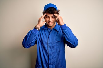 Young mechanic man wearing blue cap and uniform standing over isolated white background with hand on head for pain in head because stress. Suffering migraine.