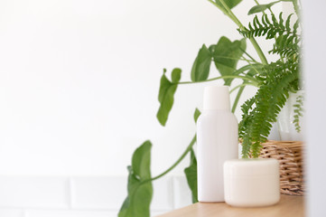 White bottle, creame jar, wicker basket with green tropic flowers over white wall.