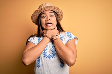 Young beautiful asian girl wearing casual t-shirt and hat standing over yellow background shouting suffocate because painful strangle. Health problem. Asphyxiate and suicide concept.