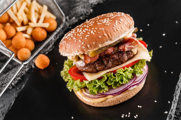 Beef burger with lettuce, tomatoes, slice of cheese, ham, pastrami, sauce and fried stick balls, french fries on slate black background, close up